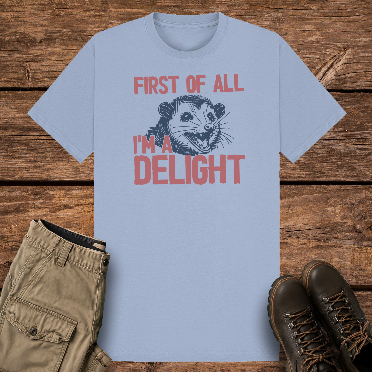 A pastel blue-colored graphic t-shirt featuring a dark blue vintage woodcut-style illustration of an opossum with its mouth wide open. Dusty pink text surrounding the image reads, "FIRST OF ALL I'M A DELIGHT."