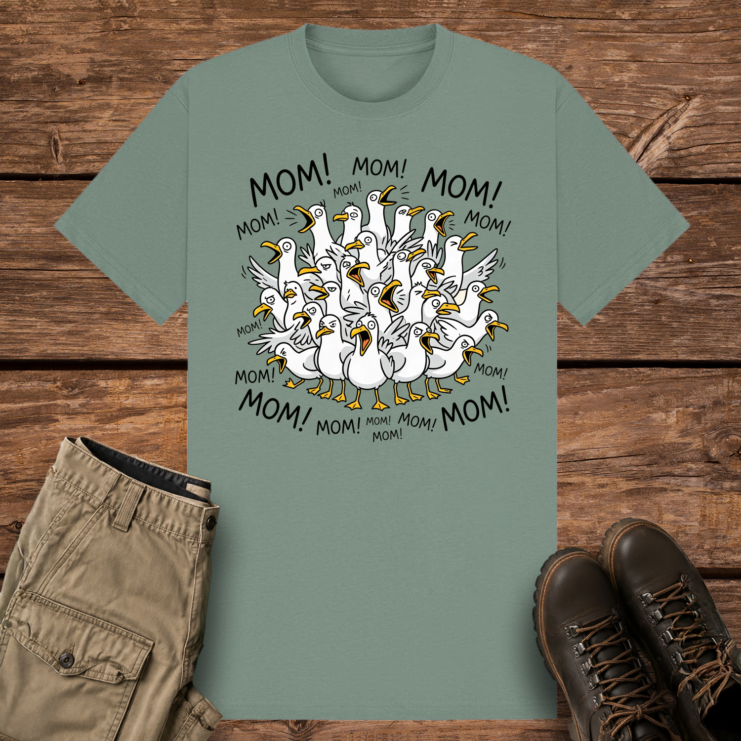 A sage-colored graphic t-shirt featuring a funny cartoon illustration of a crowded flock of squawking seagulls with their beaks wide open. The word "MOM!" is written repeatedly in a circle surrounding the frantic birds.